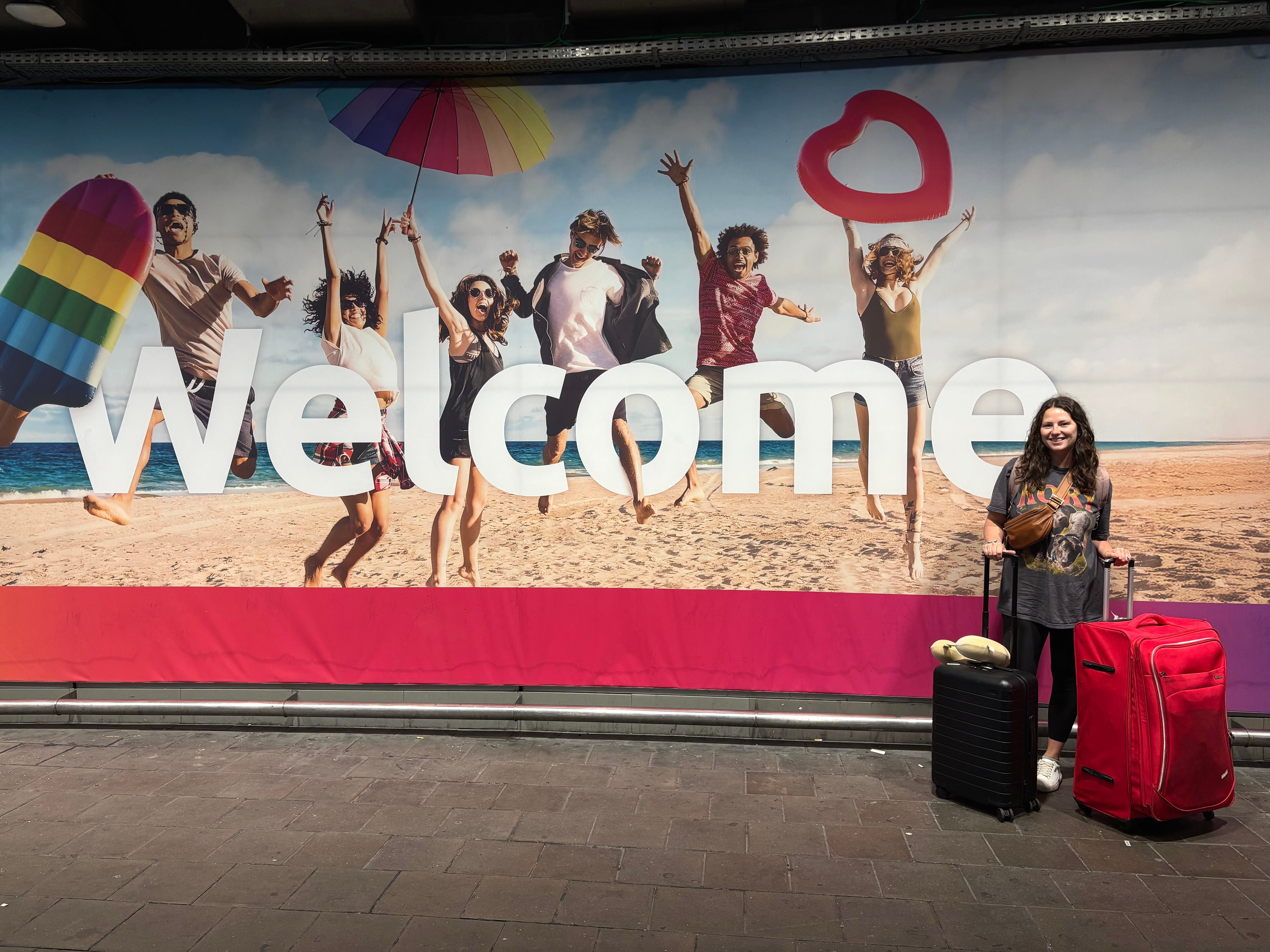 A woman stands in front of a "Welcome" sign with two suitcases.