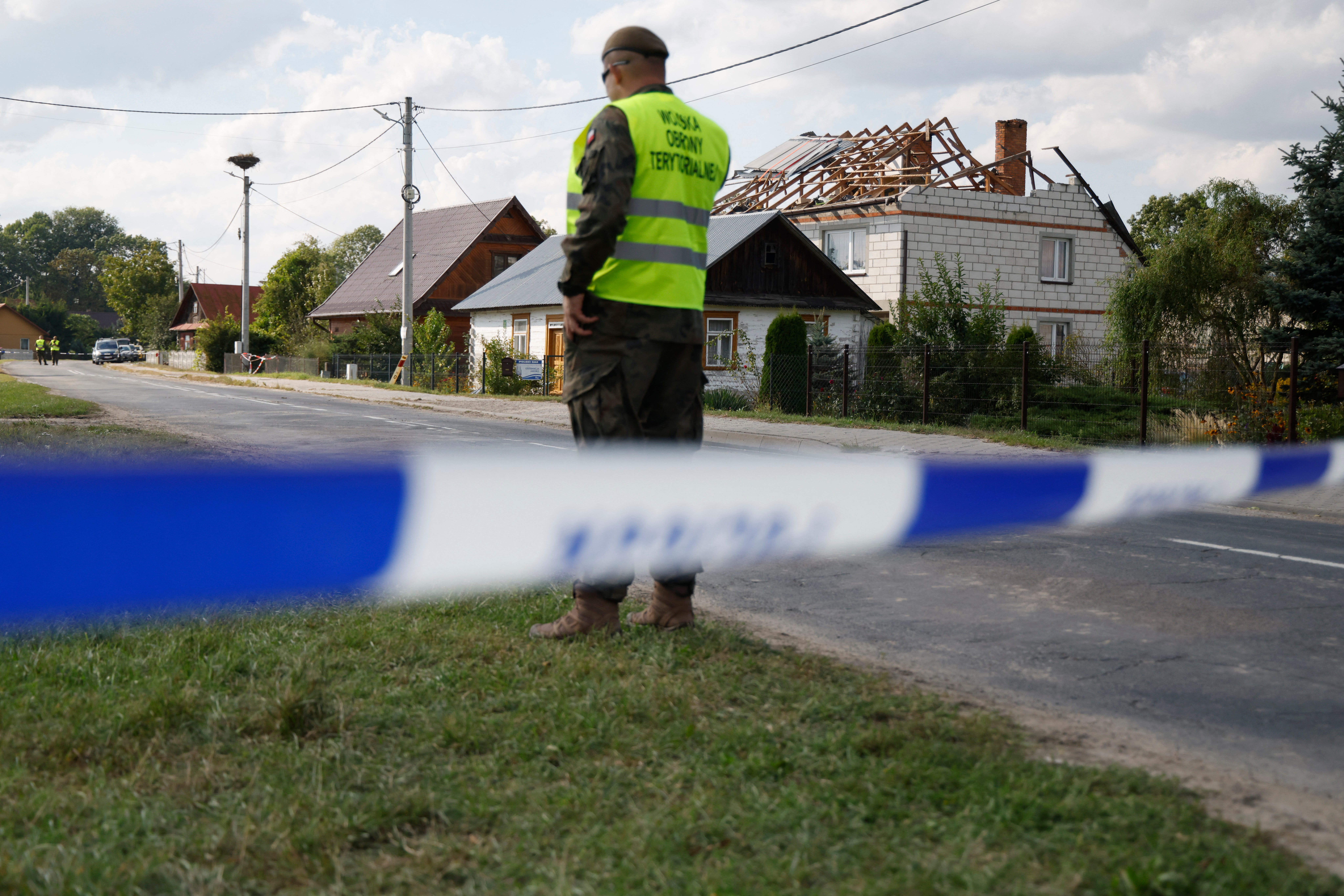 A person in a neon vest stands behind a police line in front of a house with a damaged roof