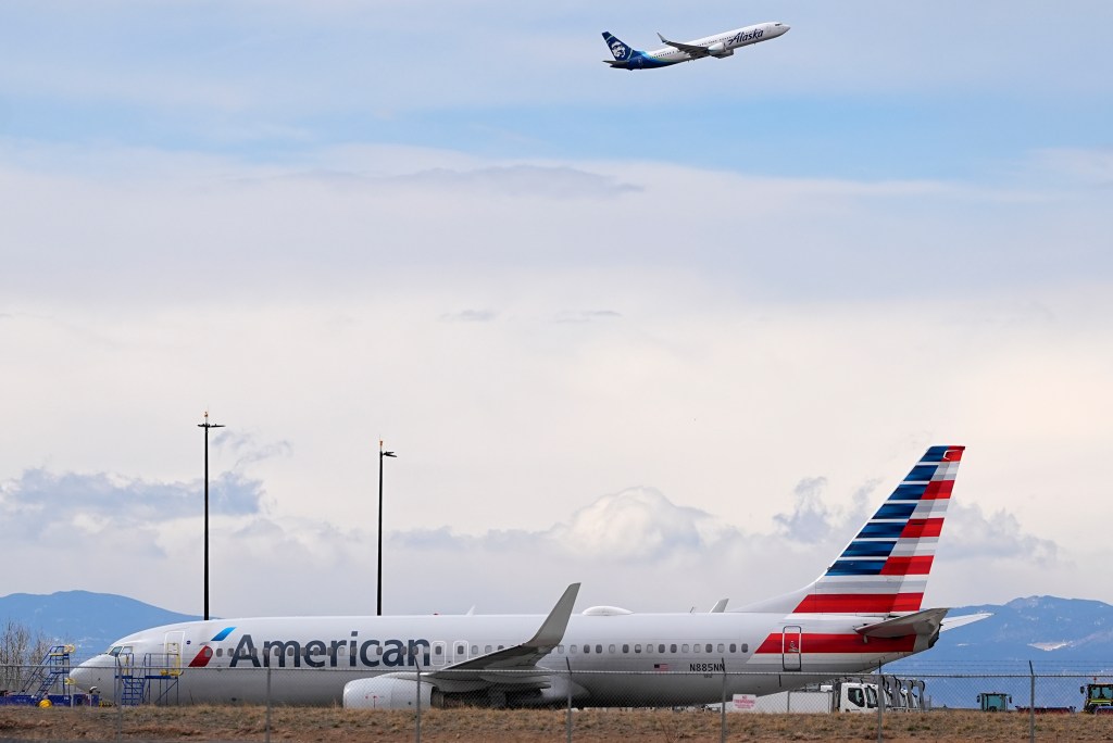An Alaska Airlines plane taking off over a grounded American Airlines plane.