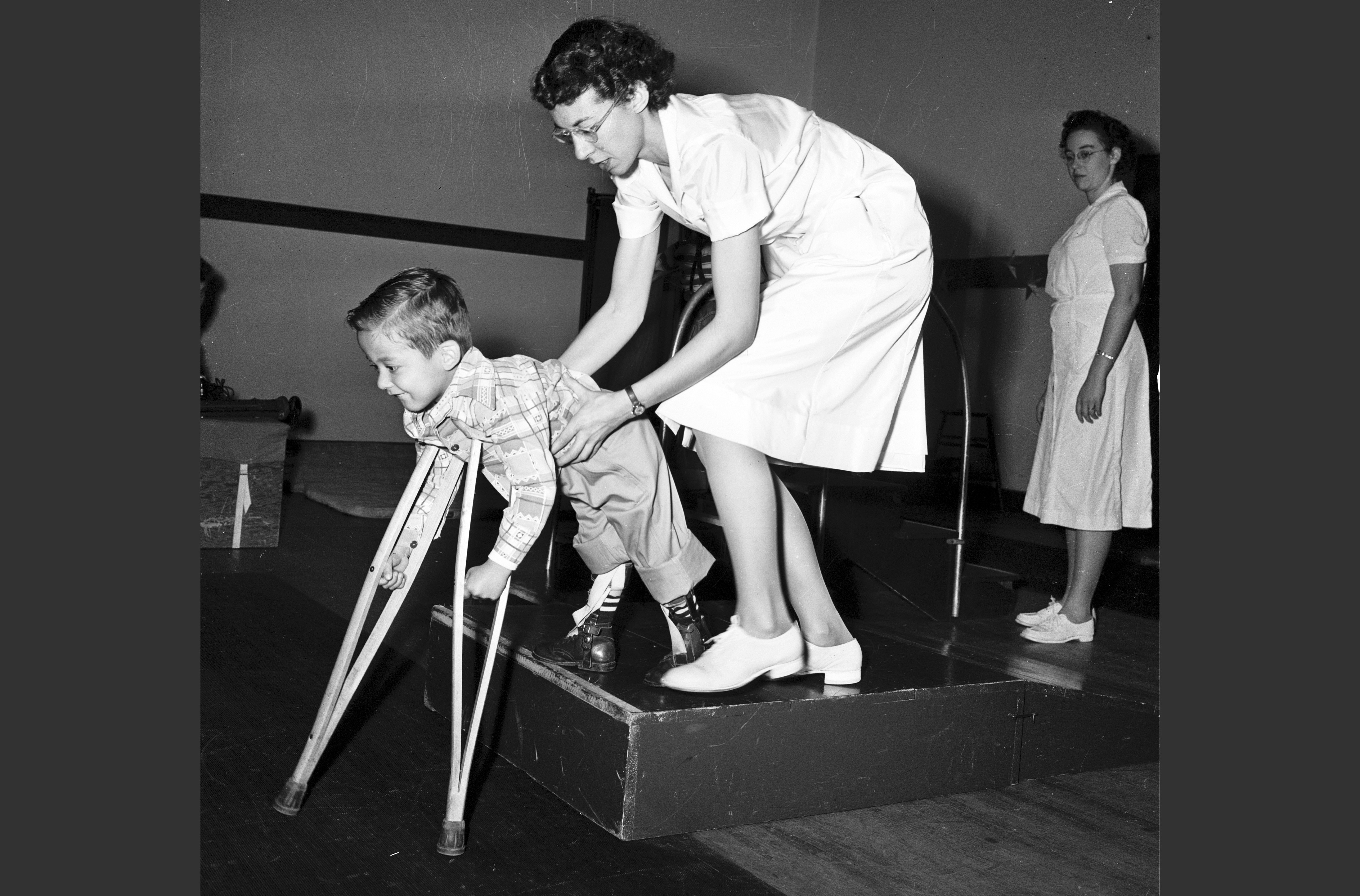 A physical therapist leans down to help as a young boy uses crutches to try and step down from a short pedestal.