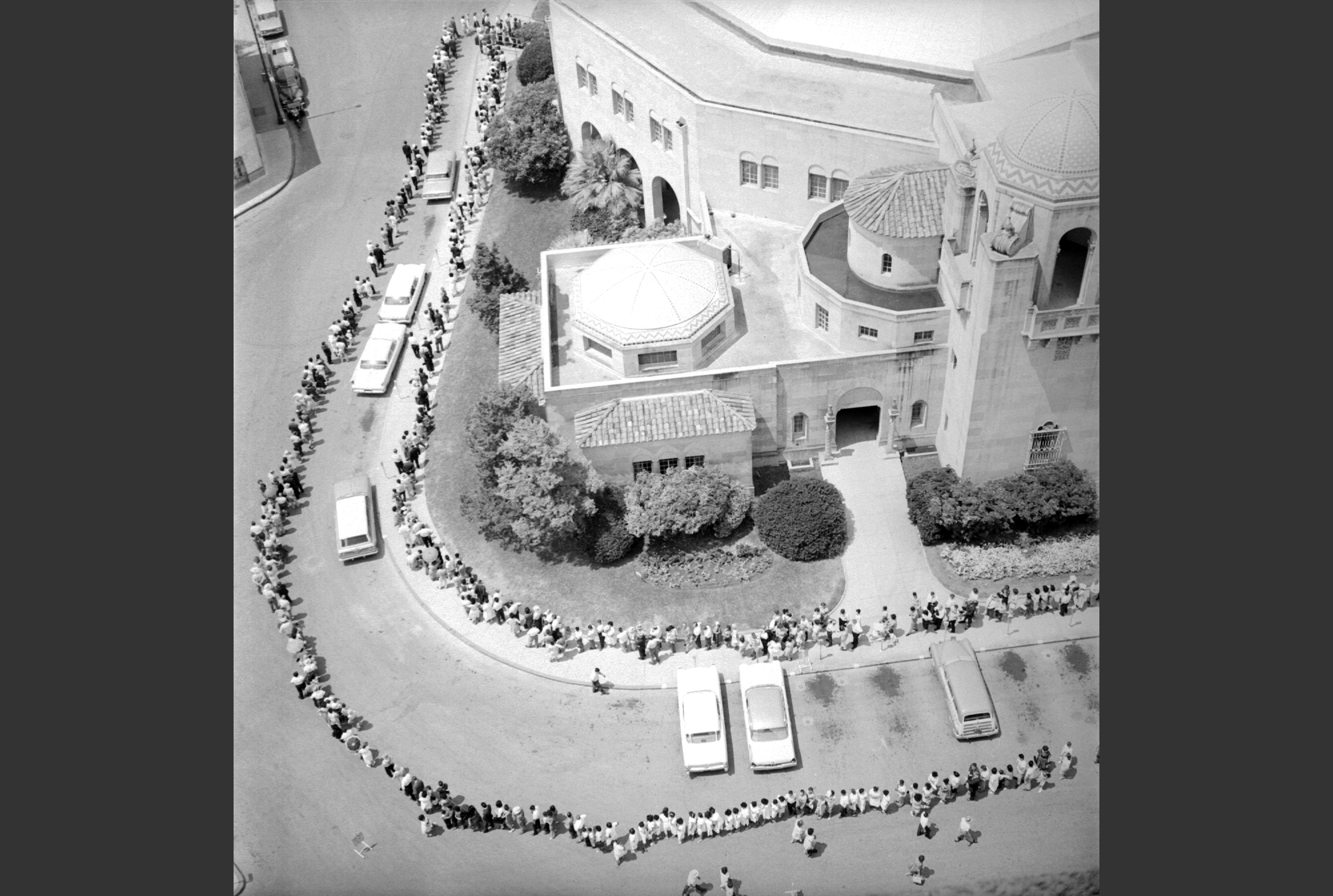 An aerial view of many people standing in long lines around the outside of an auditorium, waiting for polio vaccines.