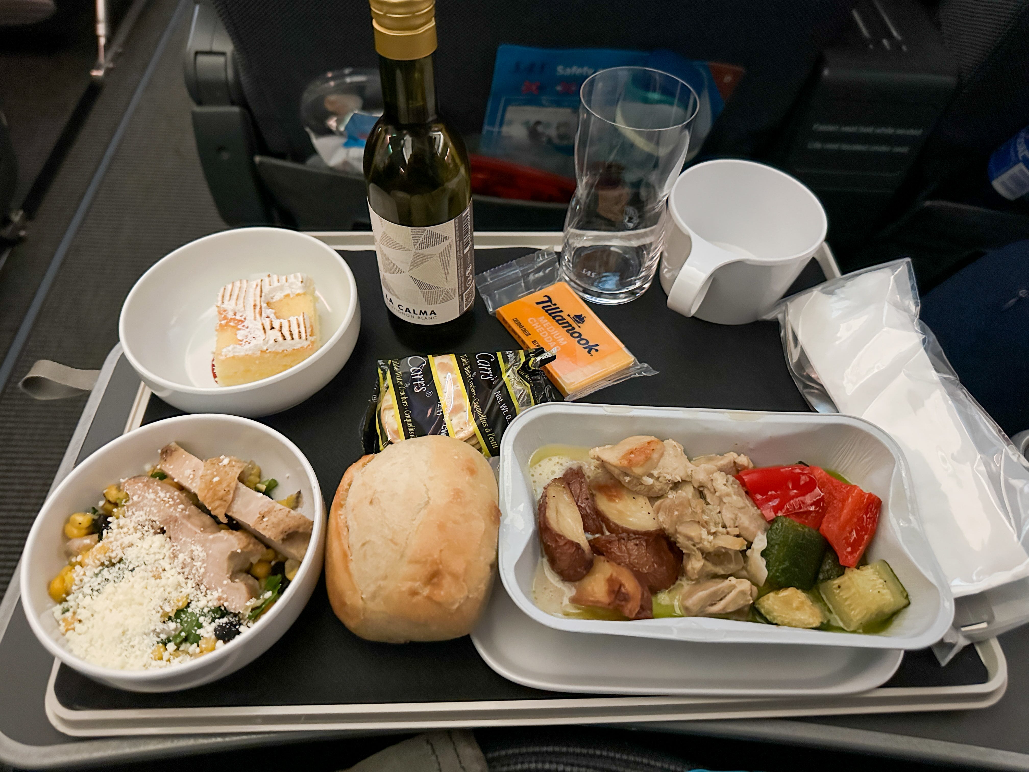 A tray of food on an airplane, including chicken with vegetables, a salad, a roll, crackers, and a dessert.