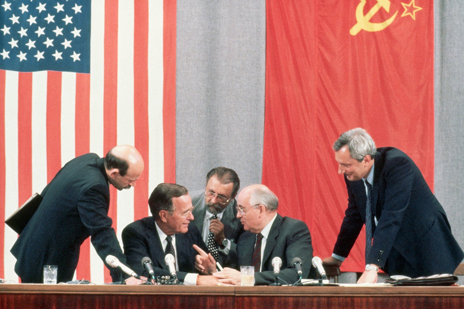 Bush and Gorbachev, both seated, lean toward each other and laugh, the latter pointing as he talks. Three other men stand and lean in toward them. The flags of the United States and the Soviet Union hang on the wall behind them.