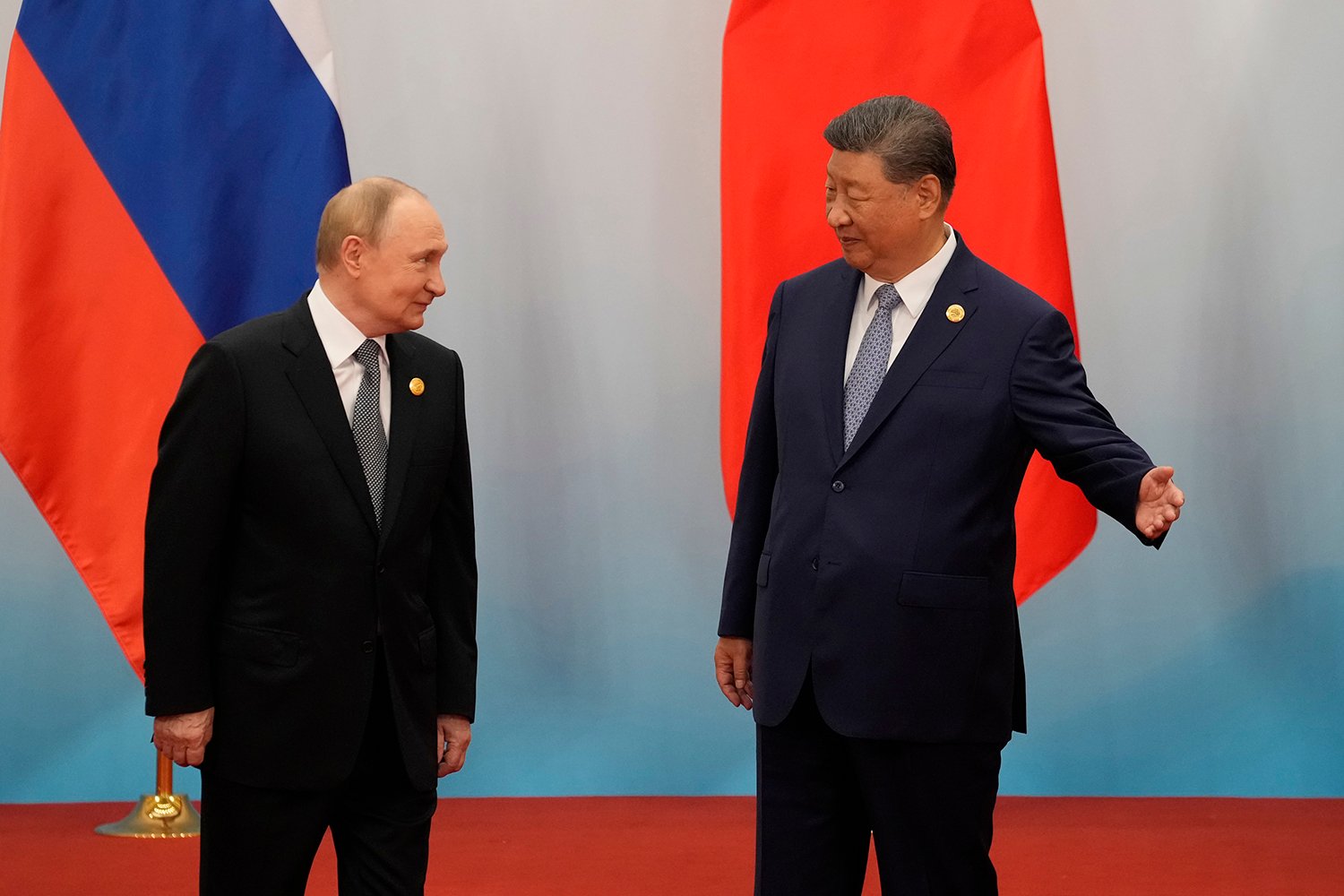 Xi Jinping gestures with his hand out as he points the way to Vladimir Putin, both in suits and standing in front of their respective countries' flags.