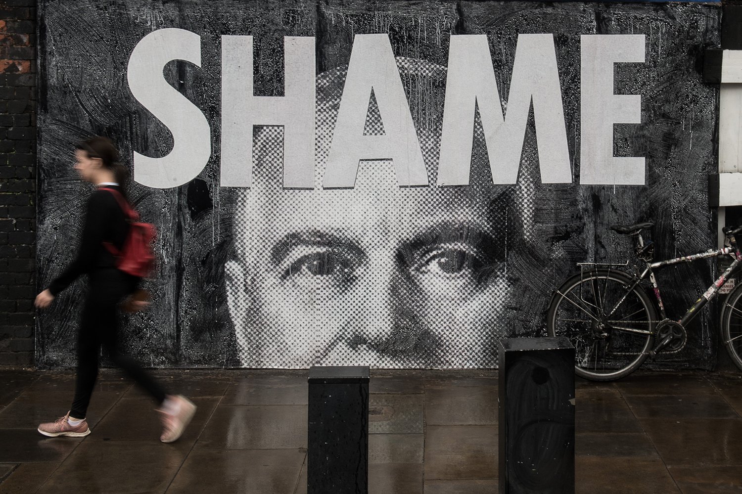 A person walks by a mural with the word SHAME over an image of the top of of Prince Andrew's face. A bike leans against the mural.