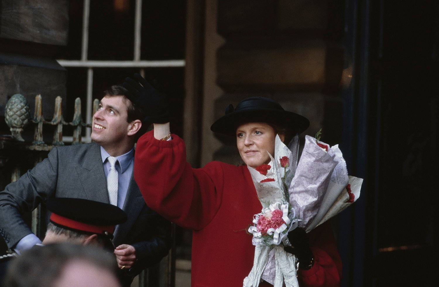 Prince Andrew looks up off camera and smiles as Sarah, wearing a black hat, red coat, and holding bouquets of flowers, waves.