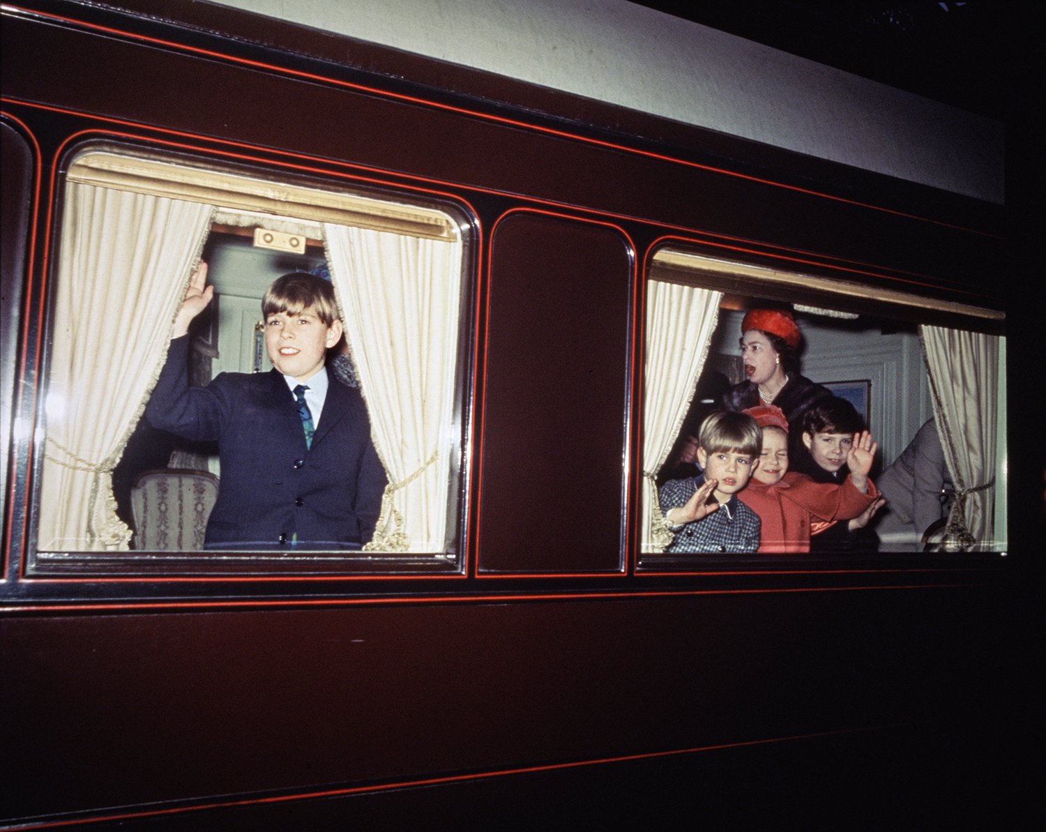 A boy waves through the curtain windows of a train. Three smaller children do the same in a rear window with the Queen behind them, her mouth agape.