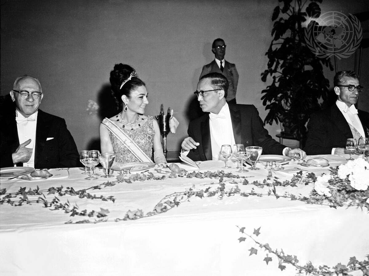 A woman in a sash, evening gown, tiara and jewels talks with a man in glasses and formal suit. Other men are on either side at a table with formal place settings and ivy vines.