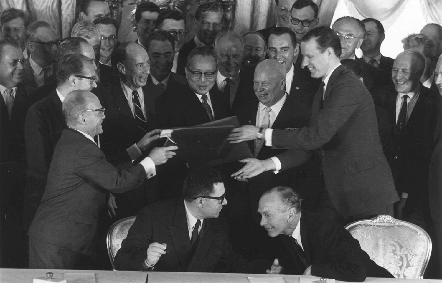 Papers are exchanged betweeen two smiling men in suits as a group of men gather around them.