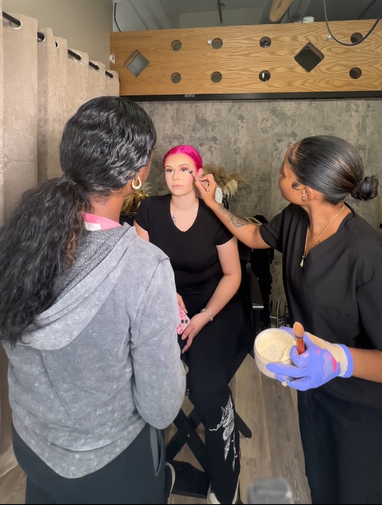 Makeup artist Alani Thompson puts makeup on a client with bright red hair as another person watches