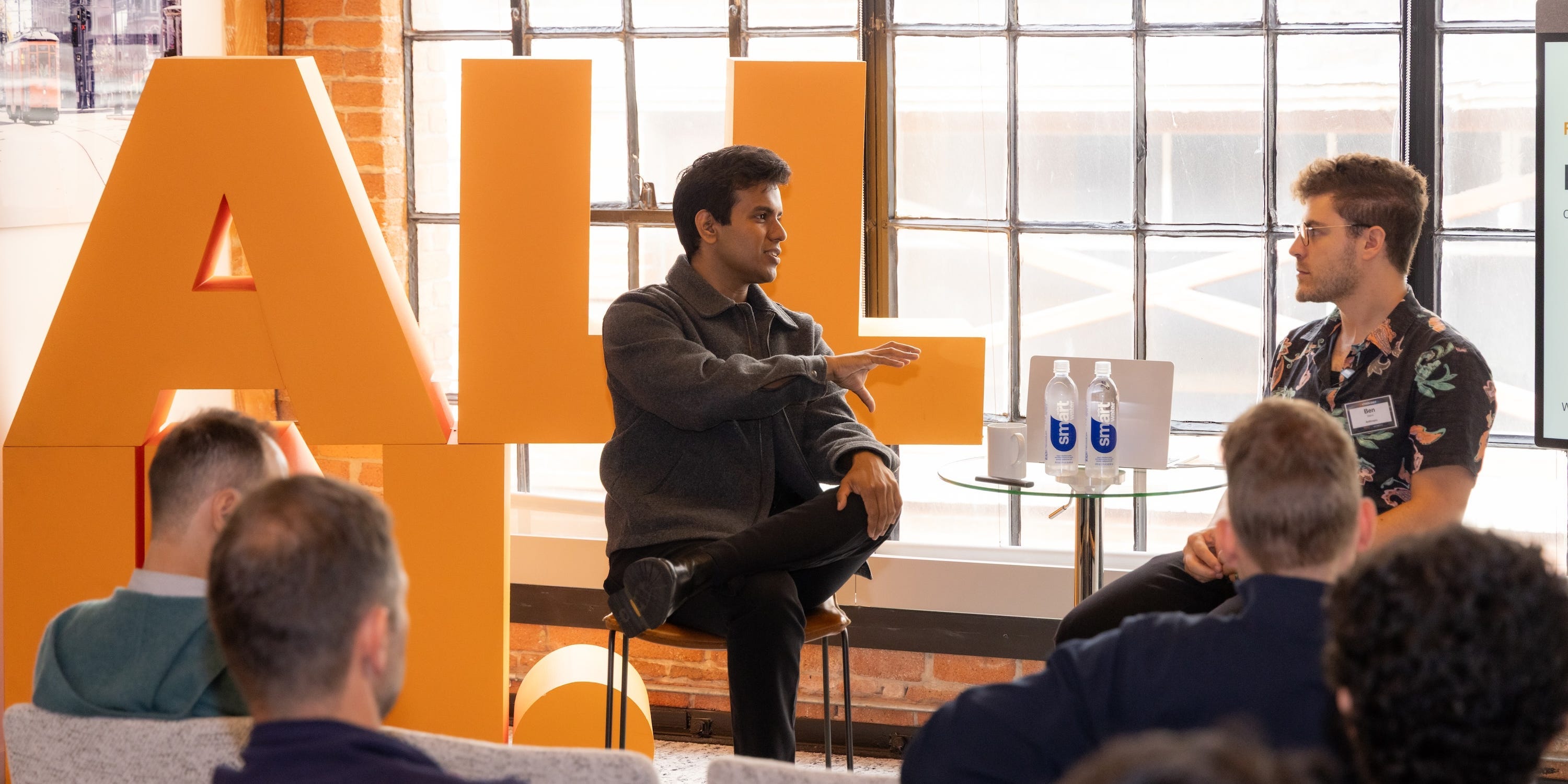 Two men talk onstage in a brightly lit workspace.