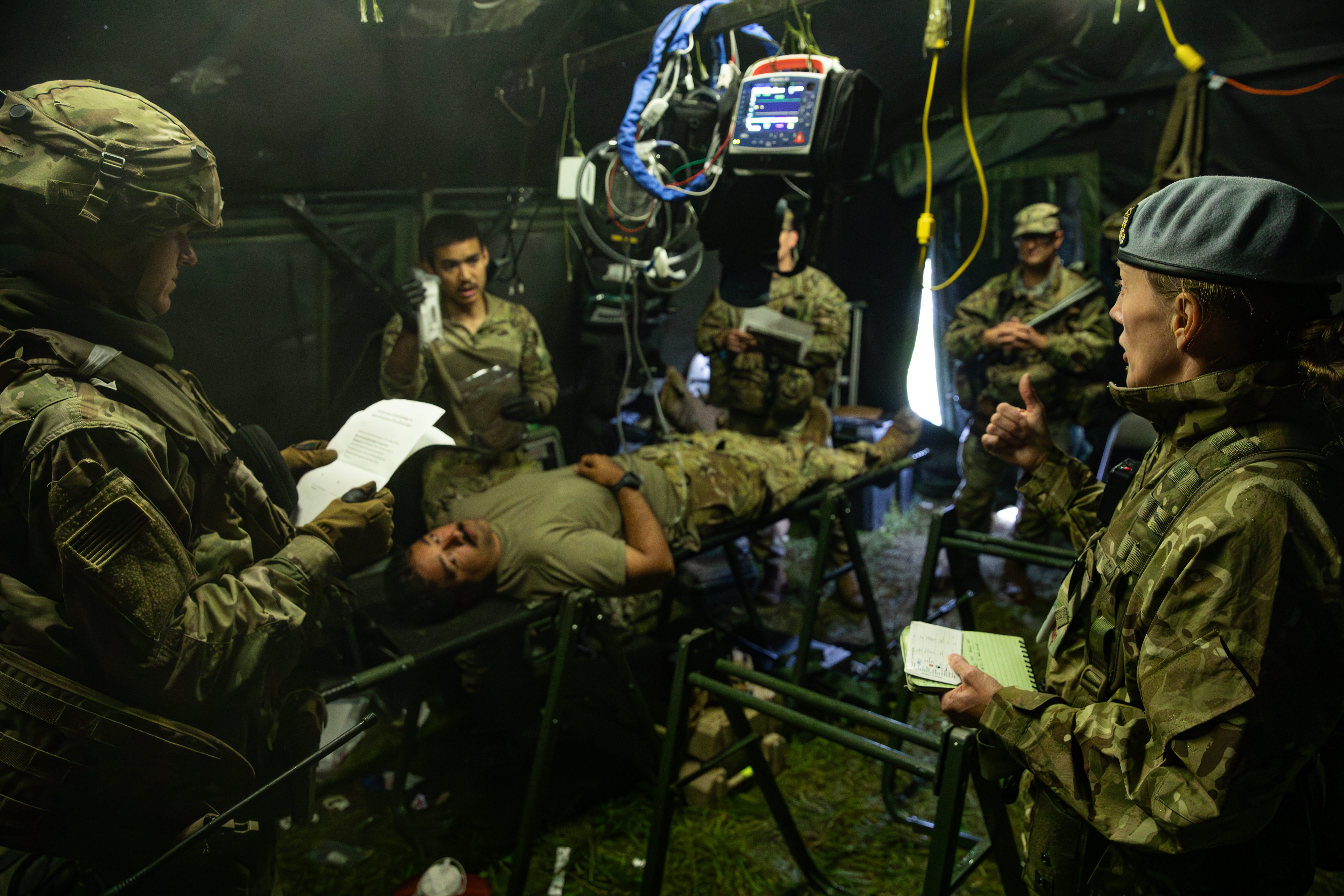 A group of military personnel wearing camouflage stand around in a medical tent. A man lies on a stretcher receiving simulated medical attention during an exercise.
