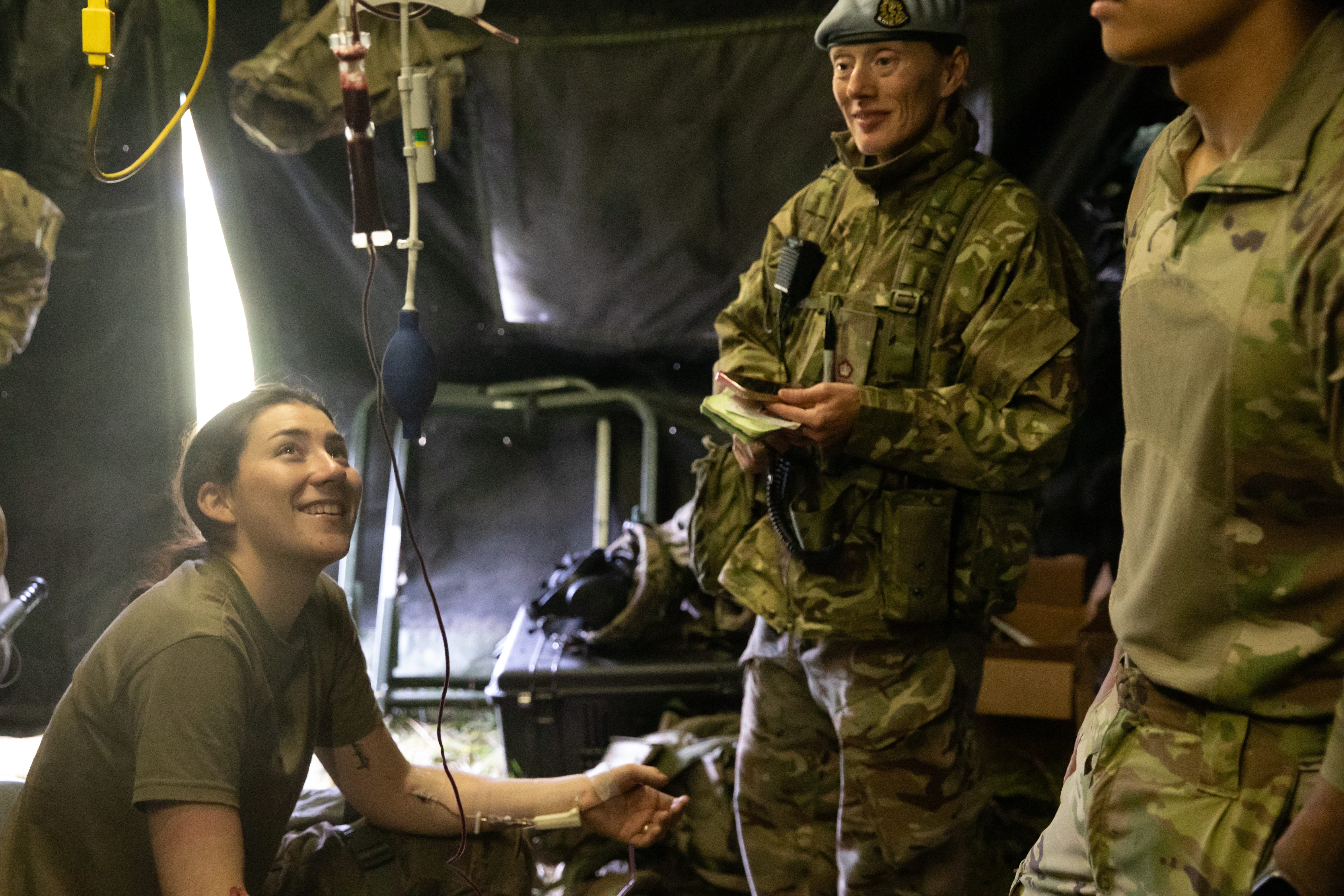 A woman wearing camouflage sits donating blood. Two people wearing camouflage stand next to her. They are all in a medical tent.