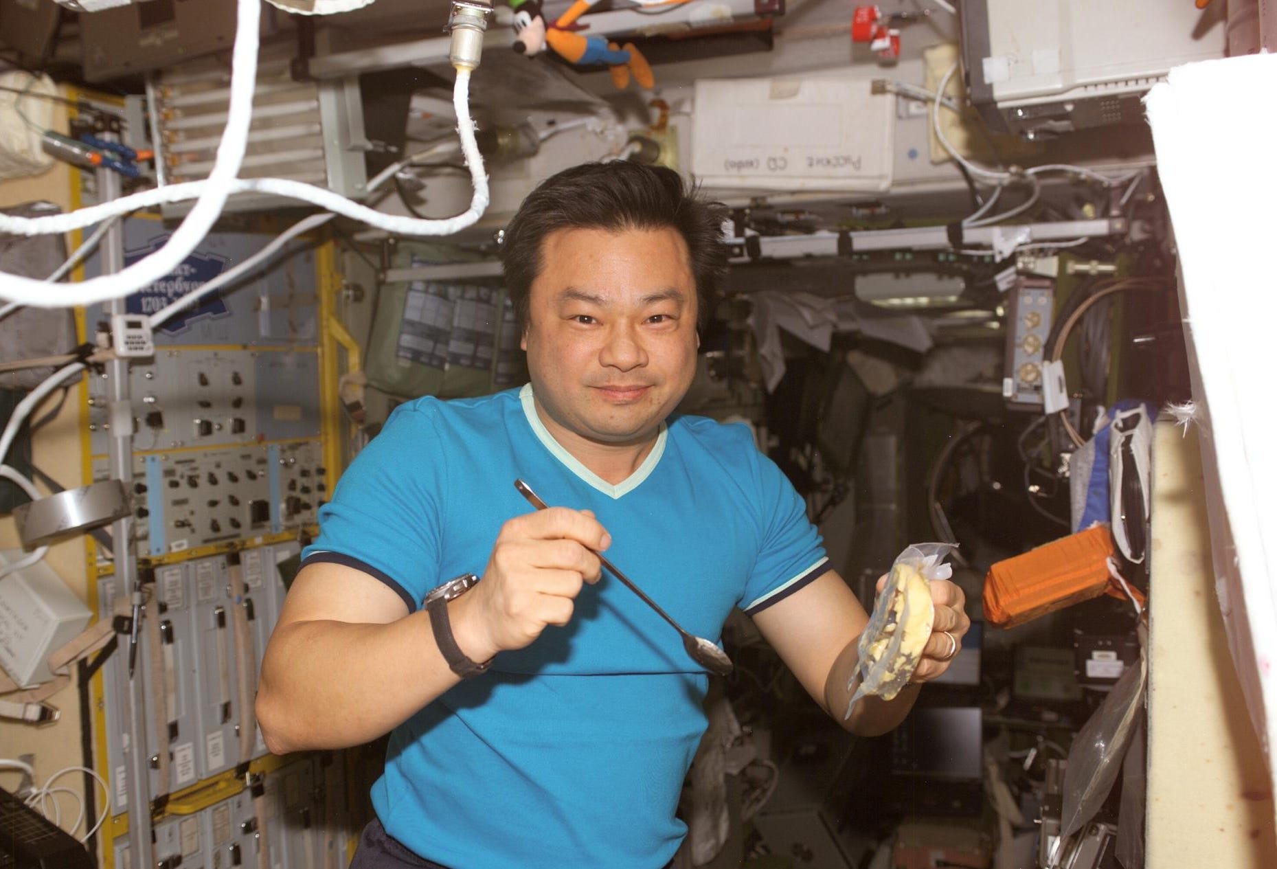 A man in a blue shirt holds a spoon and food container on the ISS