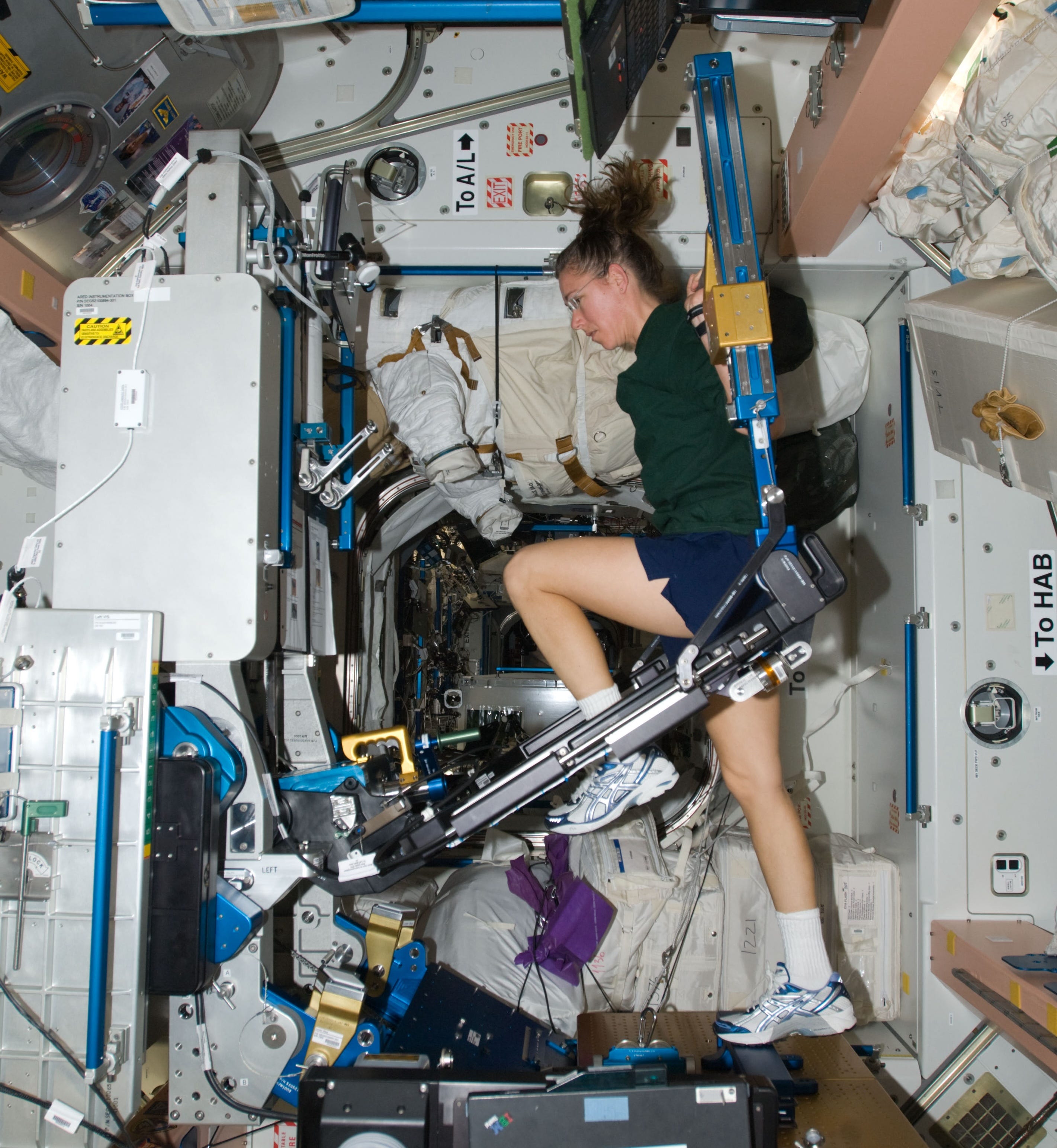 A woman in sneakers uses a large workout machine with one leg raised on the ISS