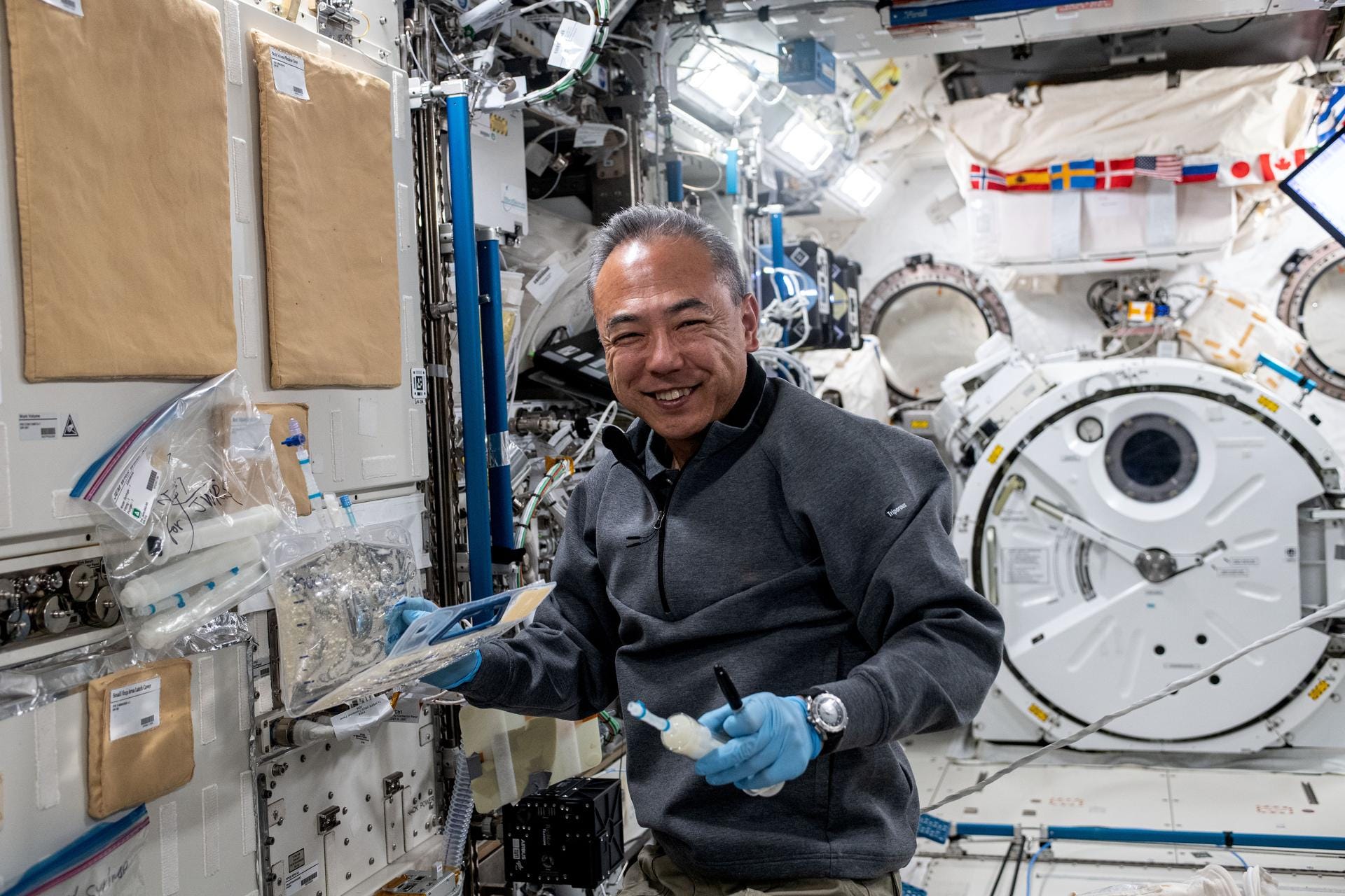 A man in a gray fleece and blue latex gloves smiles by some machinery in the ISS