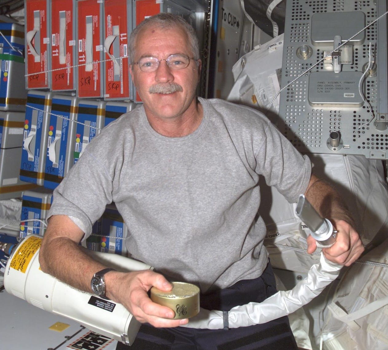 A man with a mustache and glasses holds a can and a vacuum hose on the ISS