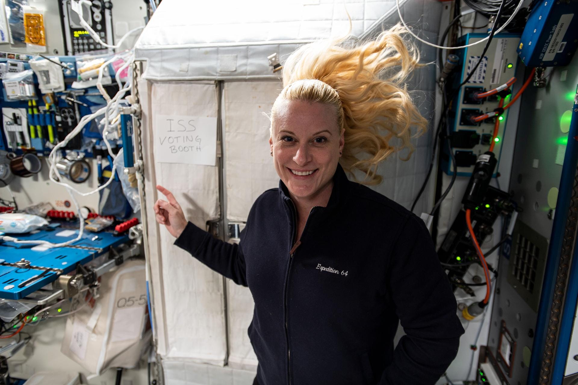 A blond woman points to a sign that says ISS voting booth on the space station