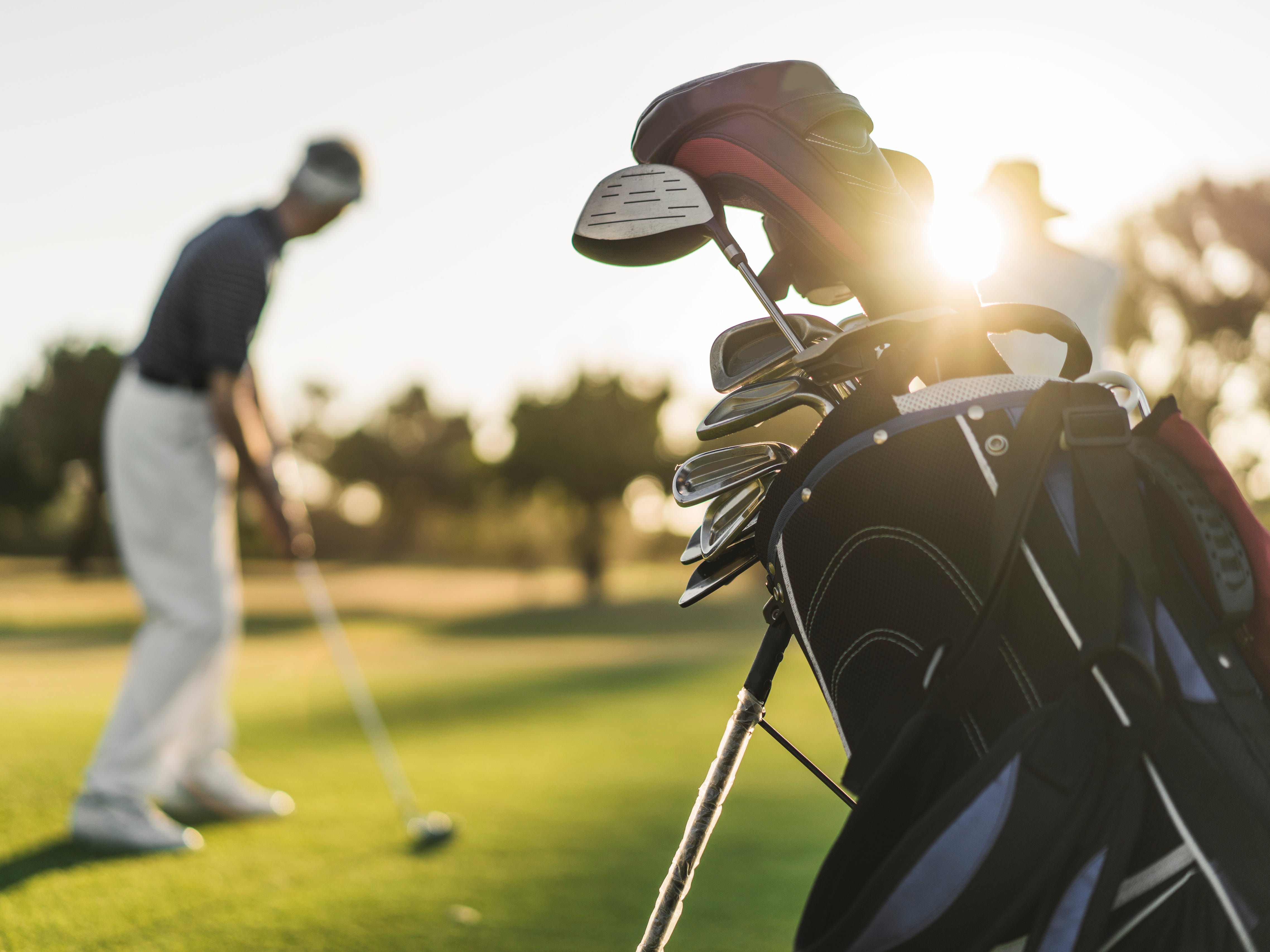A close-up of a golf bag and clubs on a course, with a man playing in the background.