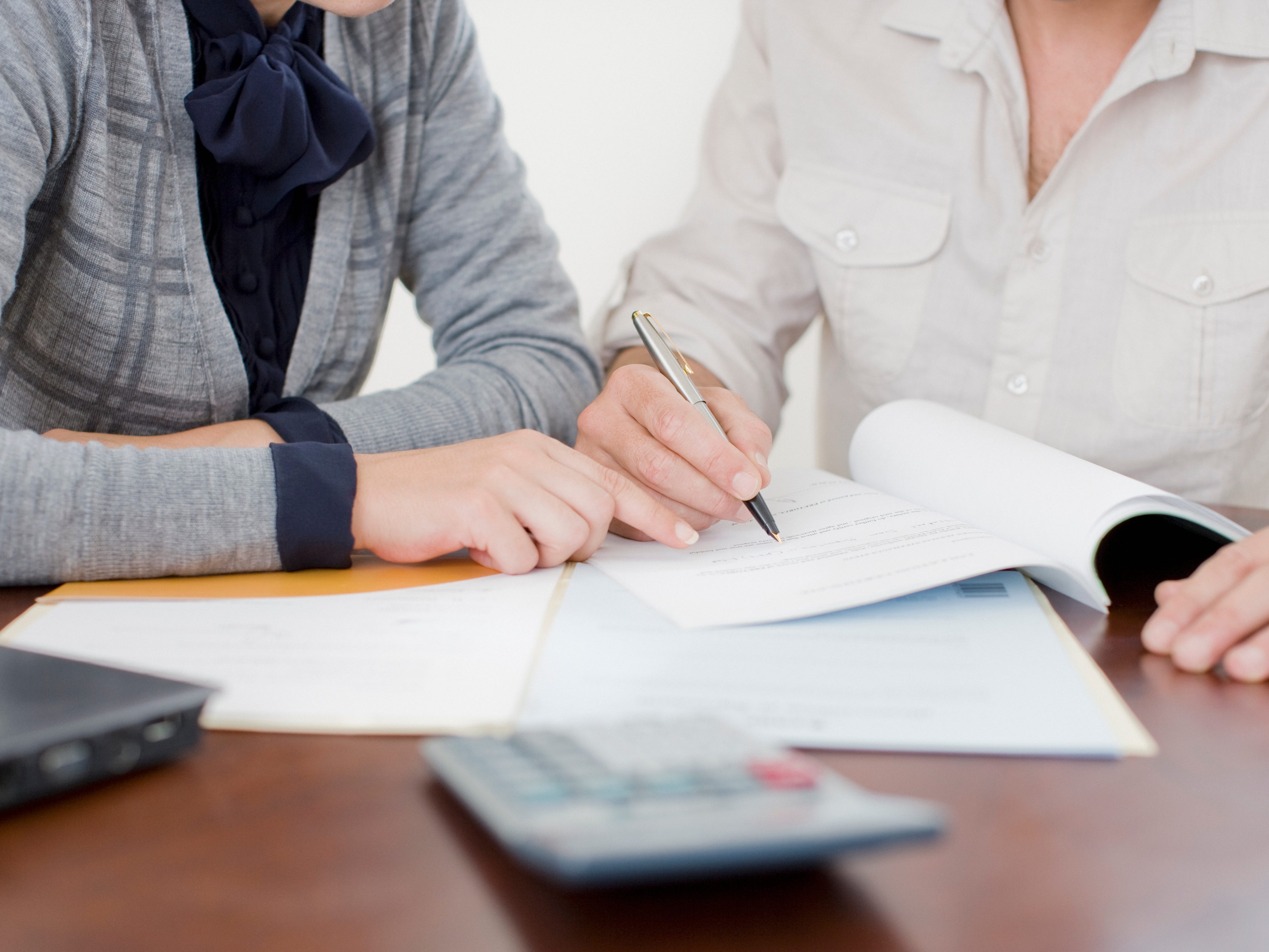 A close-up of two people on a desk, writing in a book with a calculator in front of them.