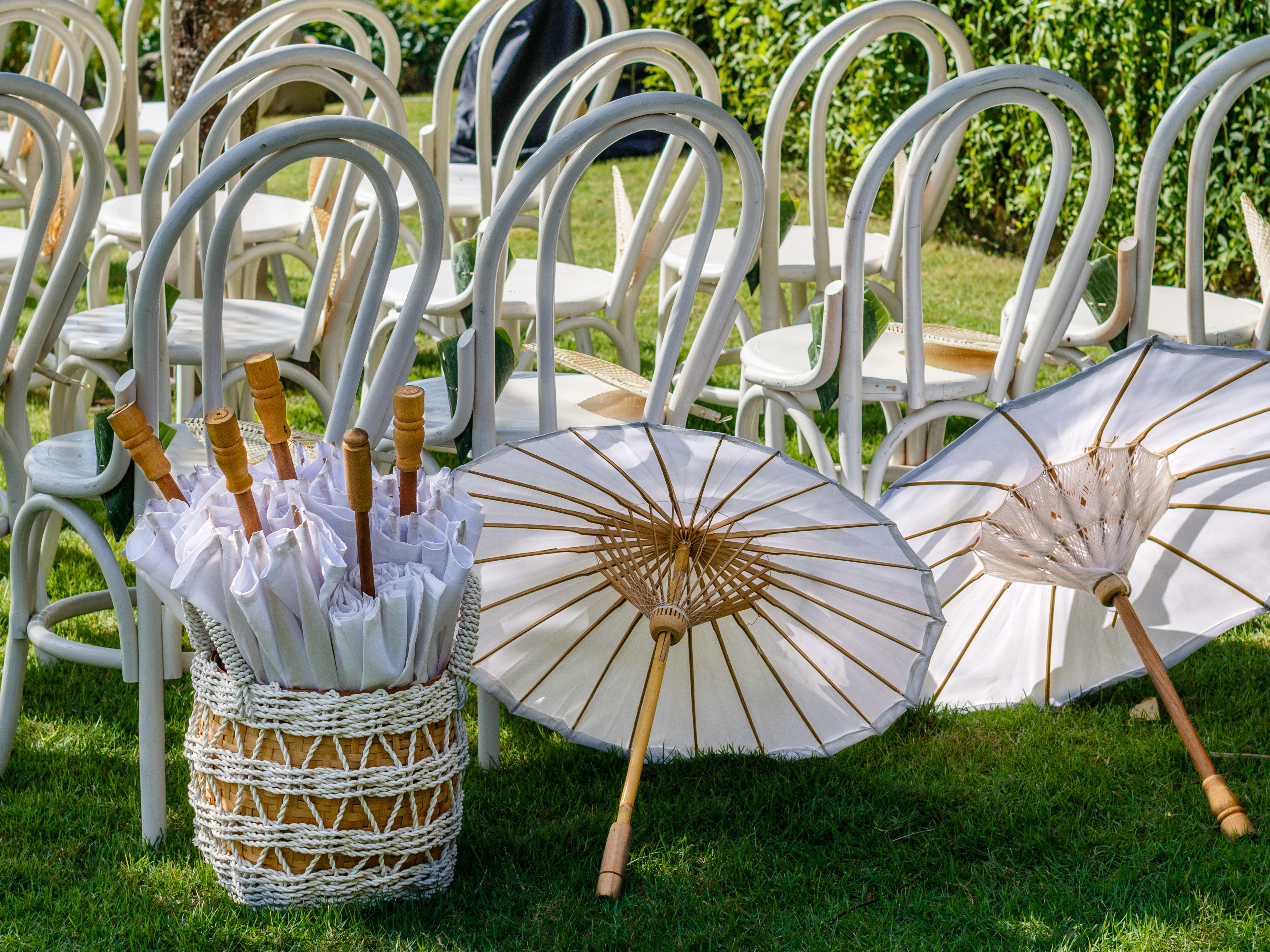 A close-up of seating at an outdoor wedding with a basket of white parasols. Two additional open parasols are on the grass nearby.