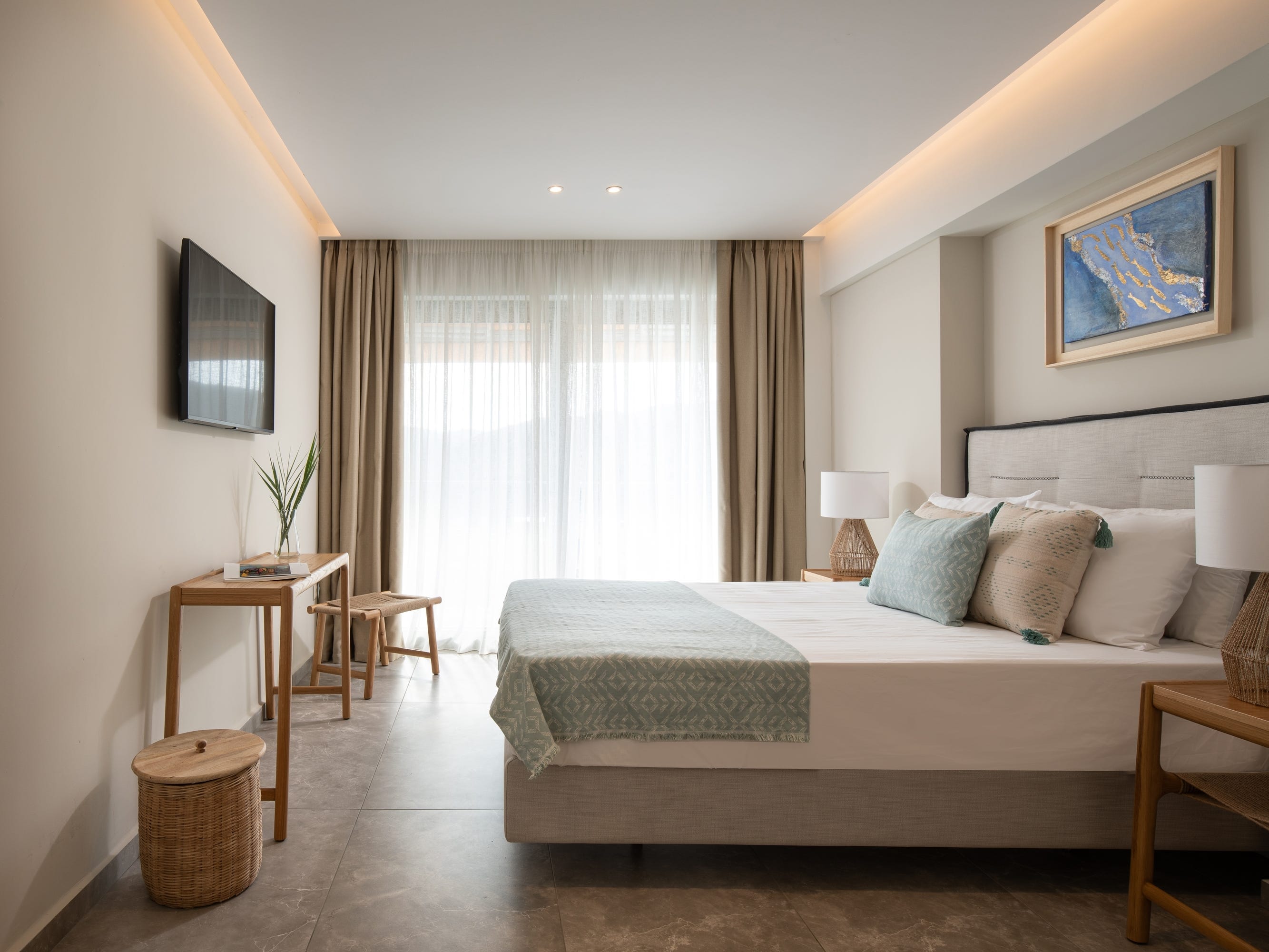 An empty hotel room, complete with a bed and a TV mounted on the wall. There's also a large window with curtains.
