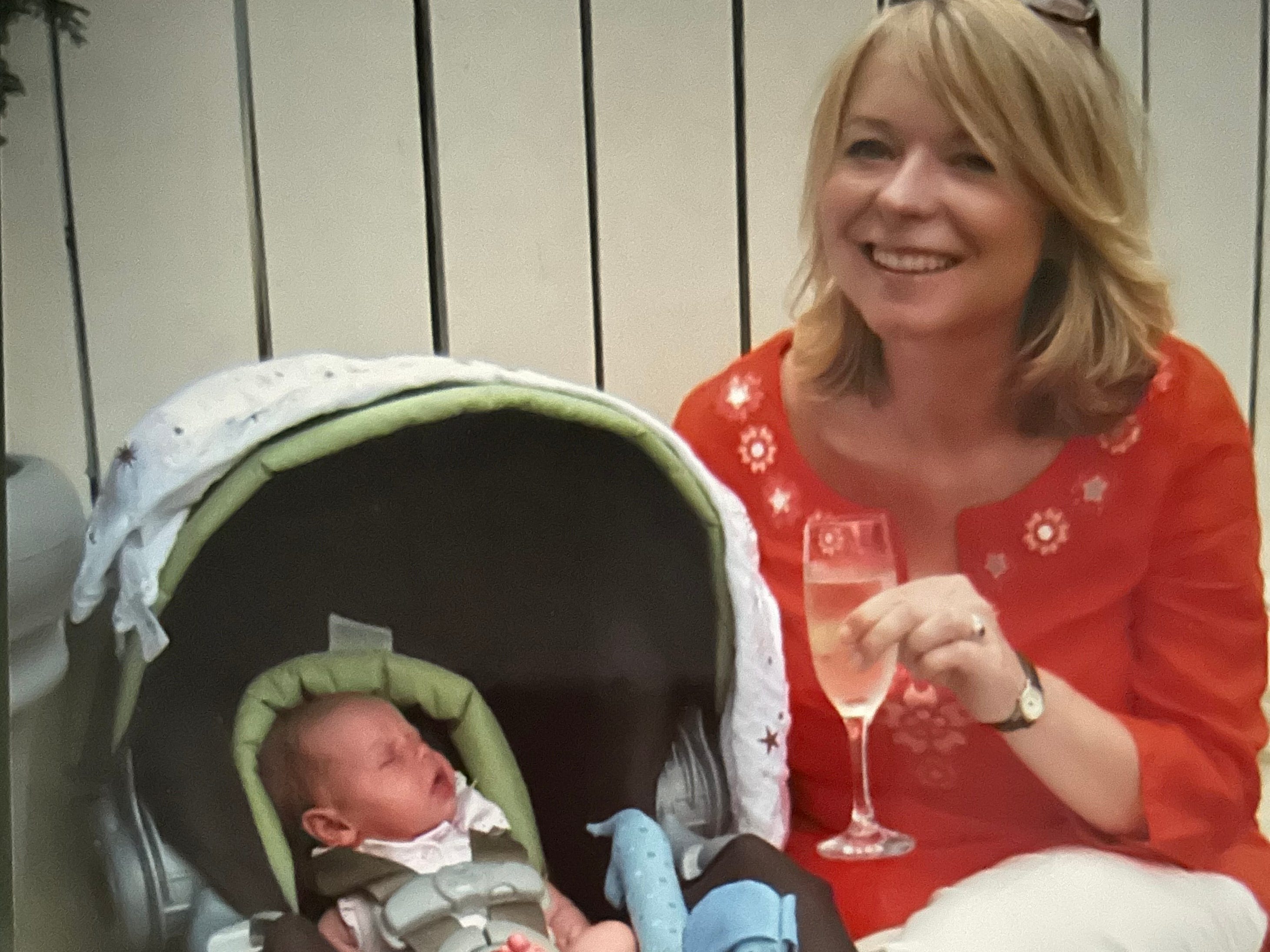 A woman with a glass of wine sitting next to a newborn in a car seat