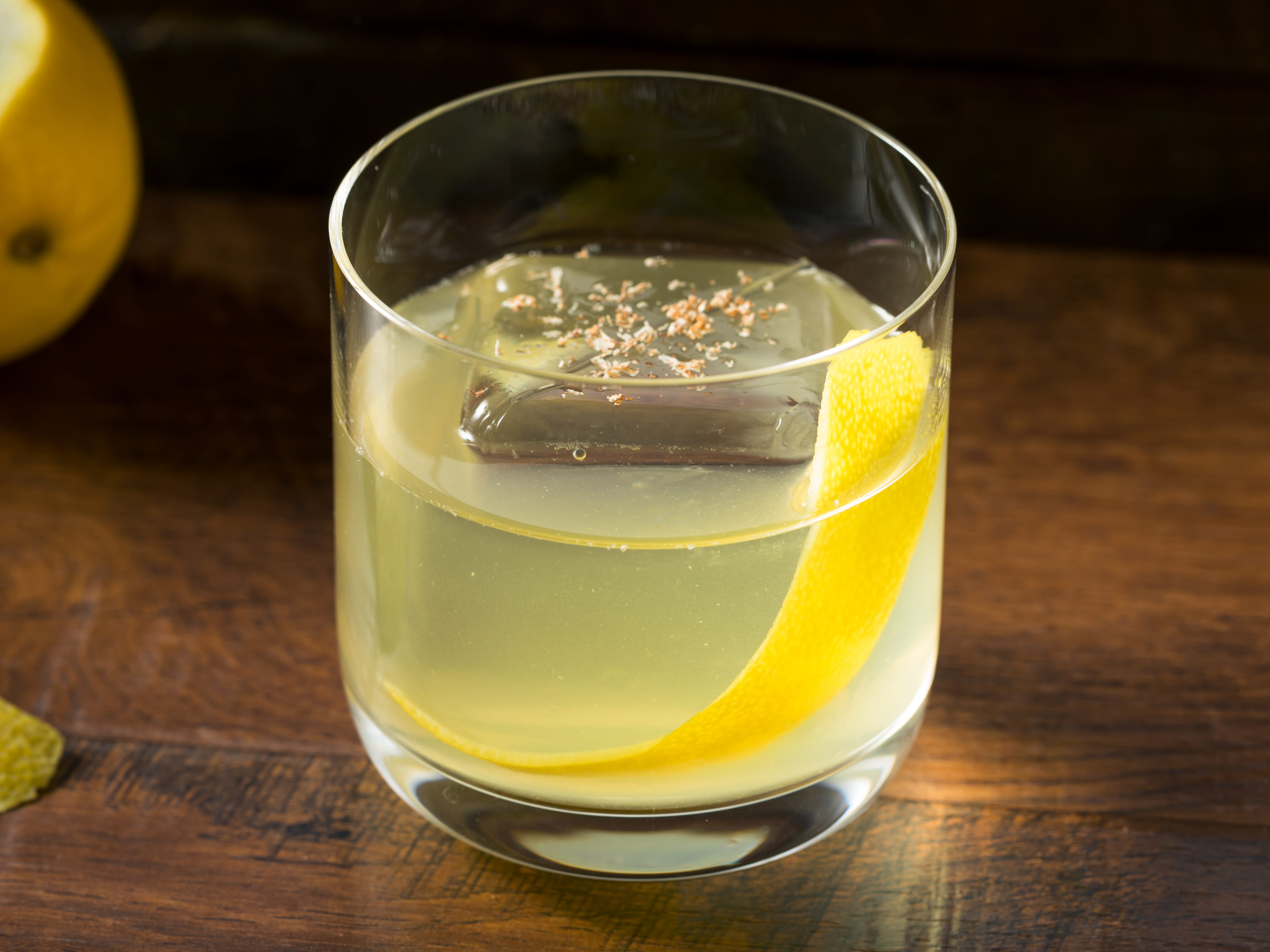 A cloudy yellow cocktail with a lemon peel and a large ice cube.