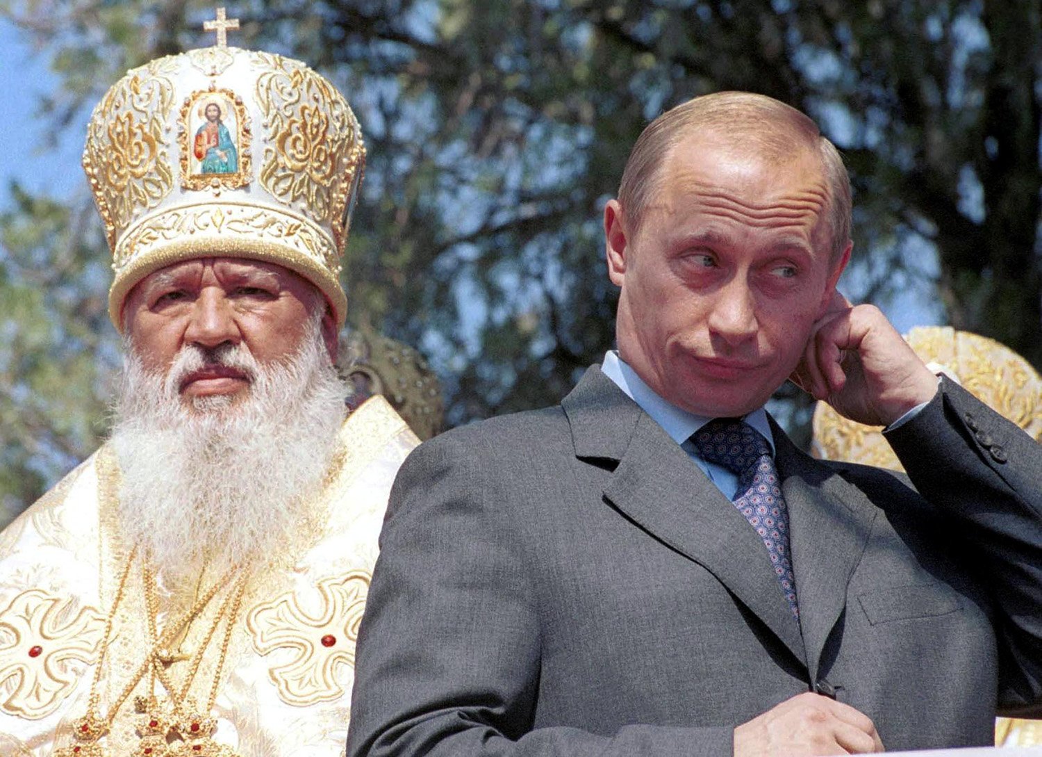 Putin, wearing a gray suit, touches his ear and squints slightly as he stands in front of a white-bearded church official in clerical robes and headwear. The branches of trees are visible behind the men.