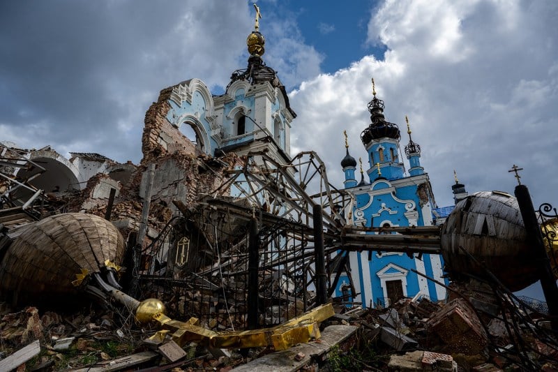 A partially destroyed church with one gilded spire resting on the ground are seen from below, framed against a mostly cloudy sky where just a bit of blue peeks out.