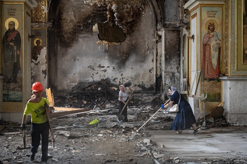 Three women push debris out of the way with brooms and their hands in a large high-ceilinged church space that shows signs of heavy damage. Orthodox-style saint icons are painted on columns on either side of the nave. One woman wears a hard hat and pants; the other two wear headscarves and skirts.
