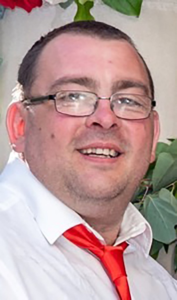 Headshot of Daniel Aitchison smiling and wearing glasses, a white shirt, and a red tie.