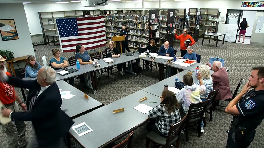 A chaotic RSU 67 school board meeting with people seated at tables, a person in an orange shirt being removed, and an American flag in the background.