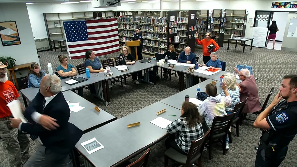 RSU 67 School Board meeting with attendees around tables and an American flag in the background.
