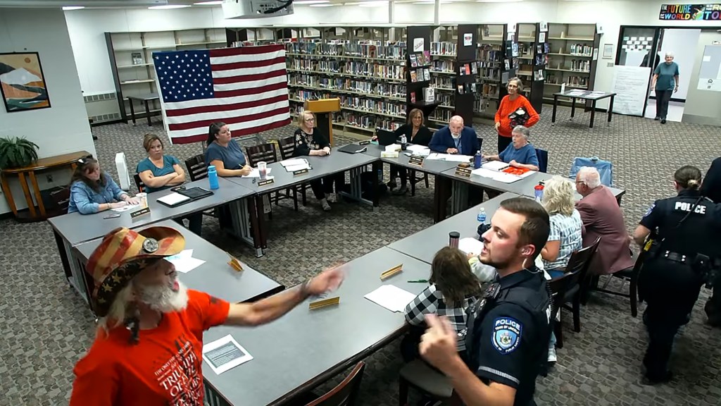 A man in a hat and an orange shirt arguing at an RSU 67 school board meeting, with police present.