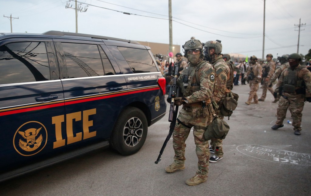 Immigration and Customs Enforcement (ICE) agents stand next to an ICE vehicle during a protest.