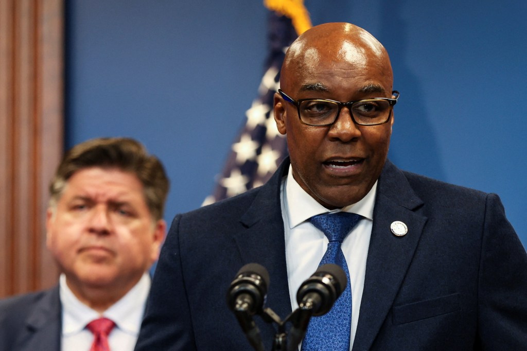 Illinois Attorney General Kwame Raoul speaking at a press conference, with Illinois Governor JB Pritzker in the background.