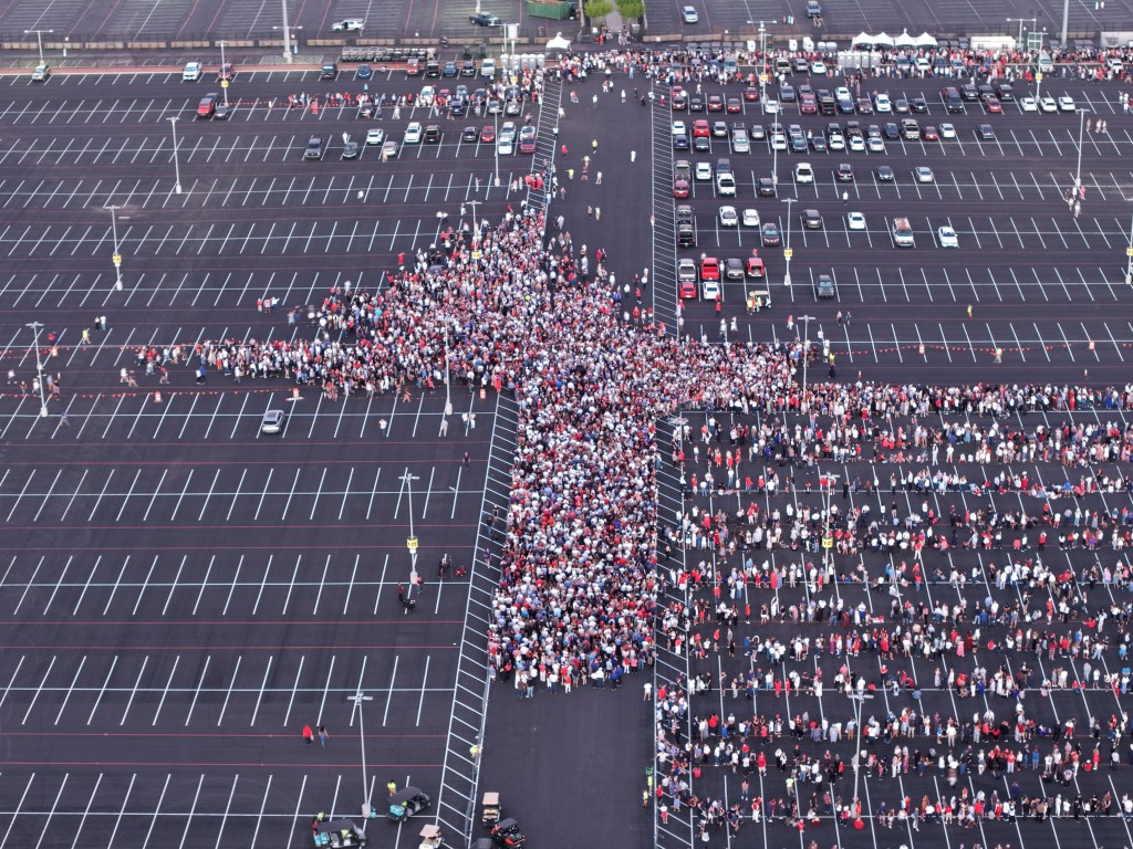 Aerial drone view of a large crowd of people, many wearing red, white, and blue, gathered in a vast parking lot for a memorial service.