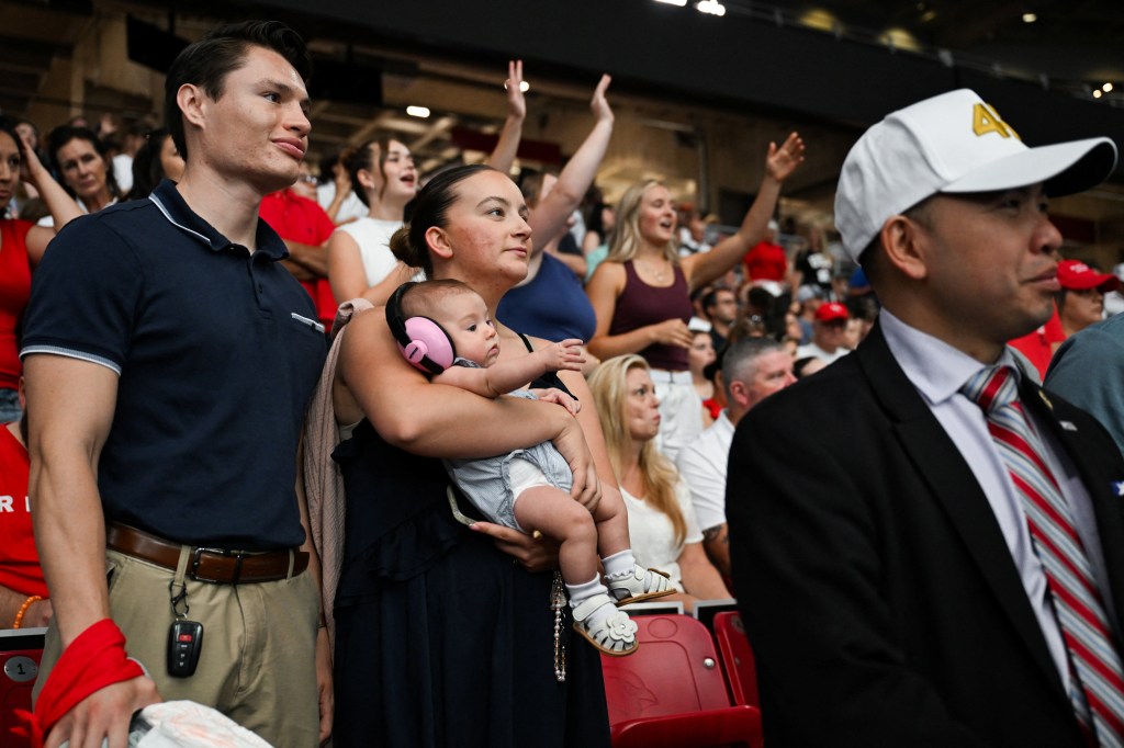 Attendees at the memorial service for Charlie Kirk, including a man in a white cap, a woman holding a baby with pink ear protection, and other people with their hands raised.