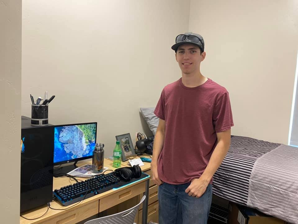 Young man in a baseball cap and t-shirt standing next to a computer setup and bed.
