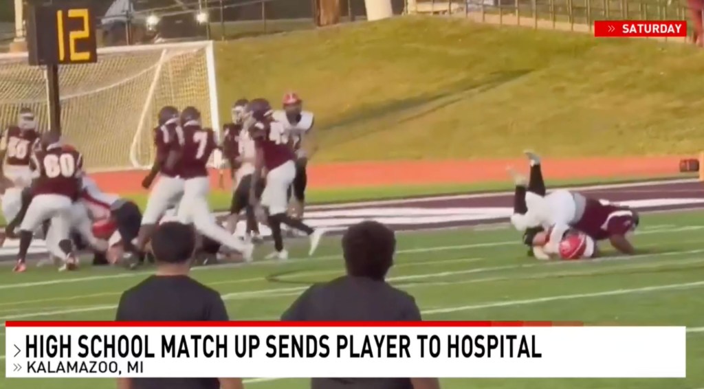 Two football players in maroon and white uniforms are on the ground after a tackle, while other players stand nearby.