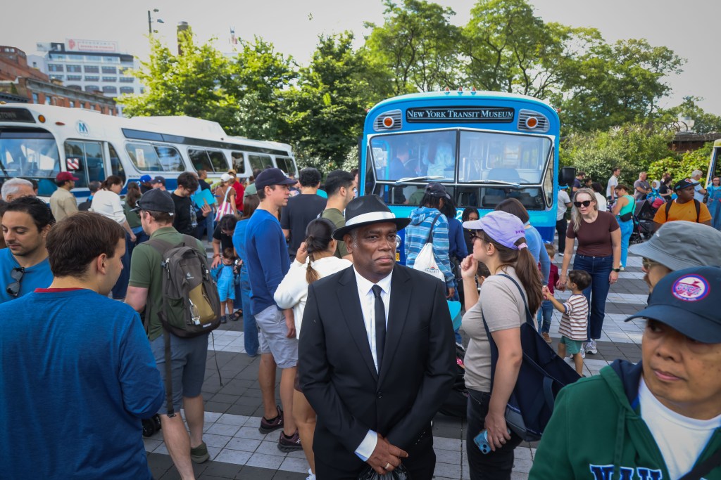A man in a suit and fedora at the Bus Festival, with buses and other attendees in the background.