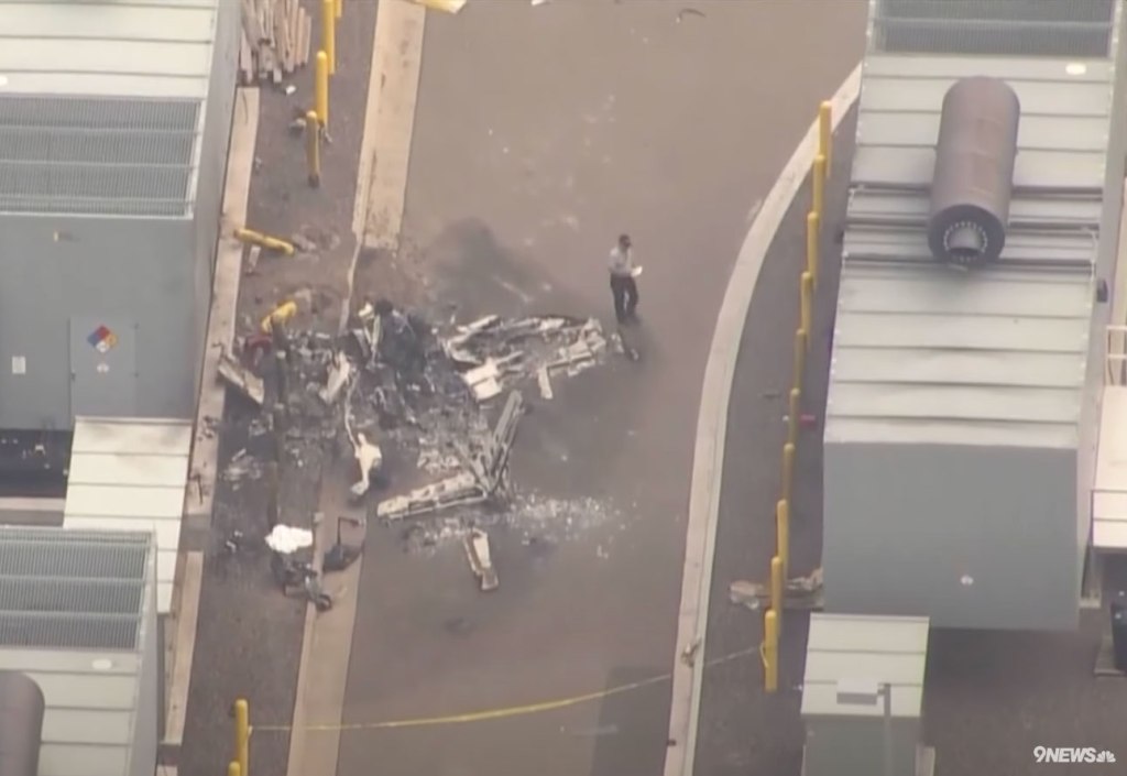 Aerial view of a helicopter crash site with debris scattered on the ground and one person standing nearby.