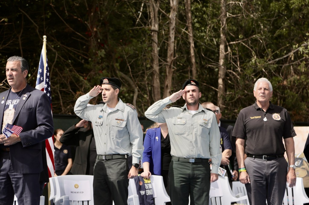 Bruce Blakeman (far right) at a park dedication, with two men in military uniforms saluting, and an American flag.