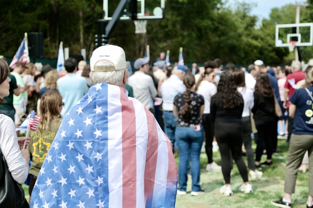 A person wrapped in an American flag, viewed from behind, in a crowd gathered in Captain Omer Neutra Memorial Park.