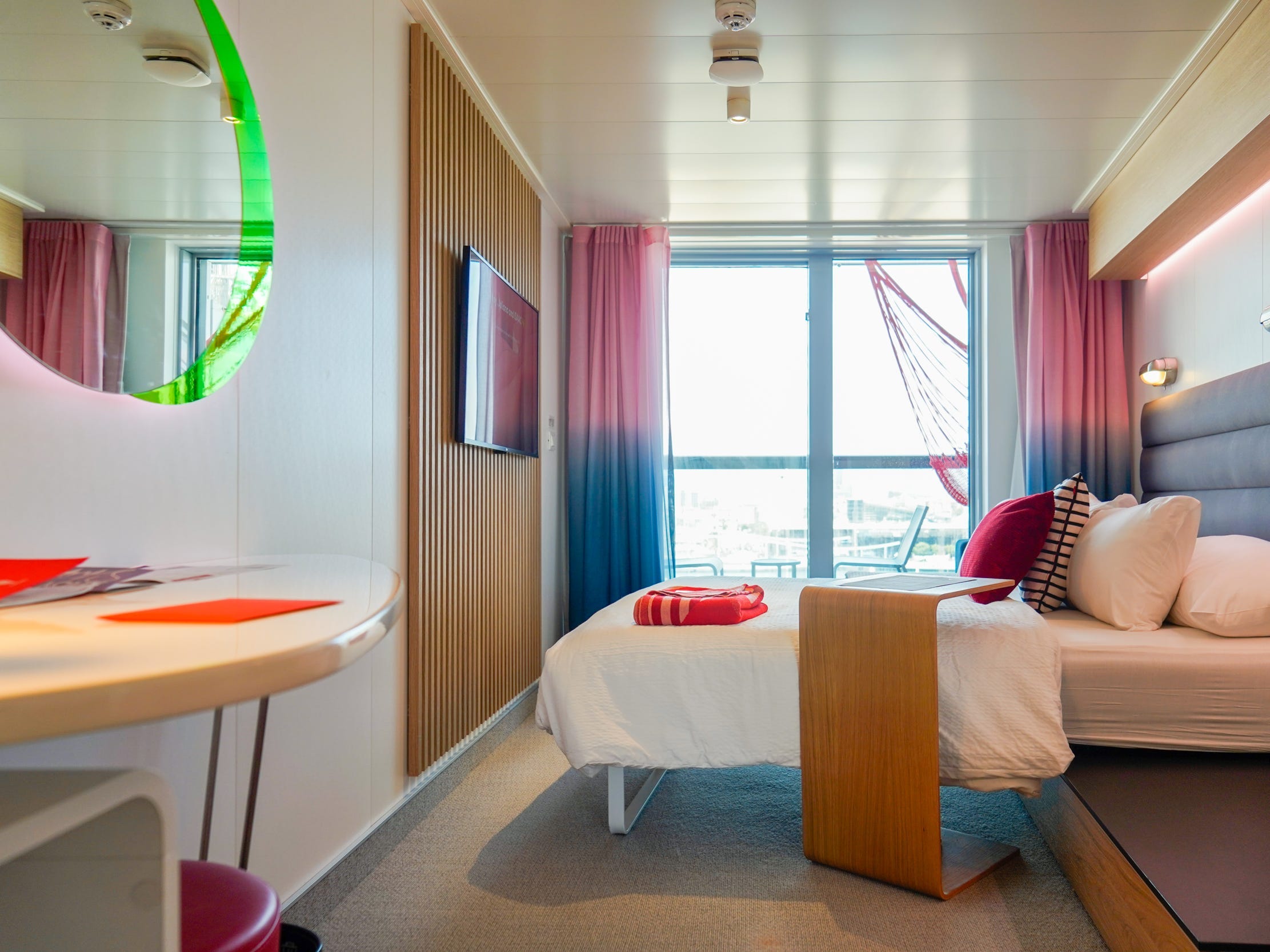 A room with a mirror and table on the left. Behind the table is a bed with a navy headboard, white sheets, and white and red pillows as well as a red bolded blanket at the bottom seen from the side in front of a large window with red and blue curtains. There's a mountain TV on a wooden-planed wall to the left  and a wooden side table next to the bed.