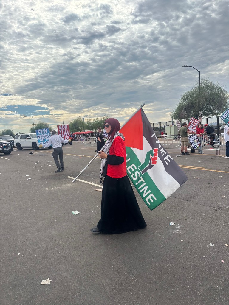 A woman wearing a red shirt, black skirt, and maroon hijab holds a Palestinian flag.