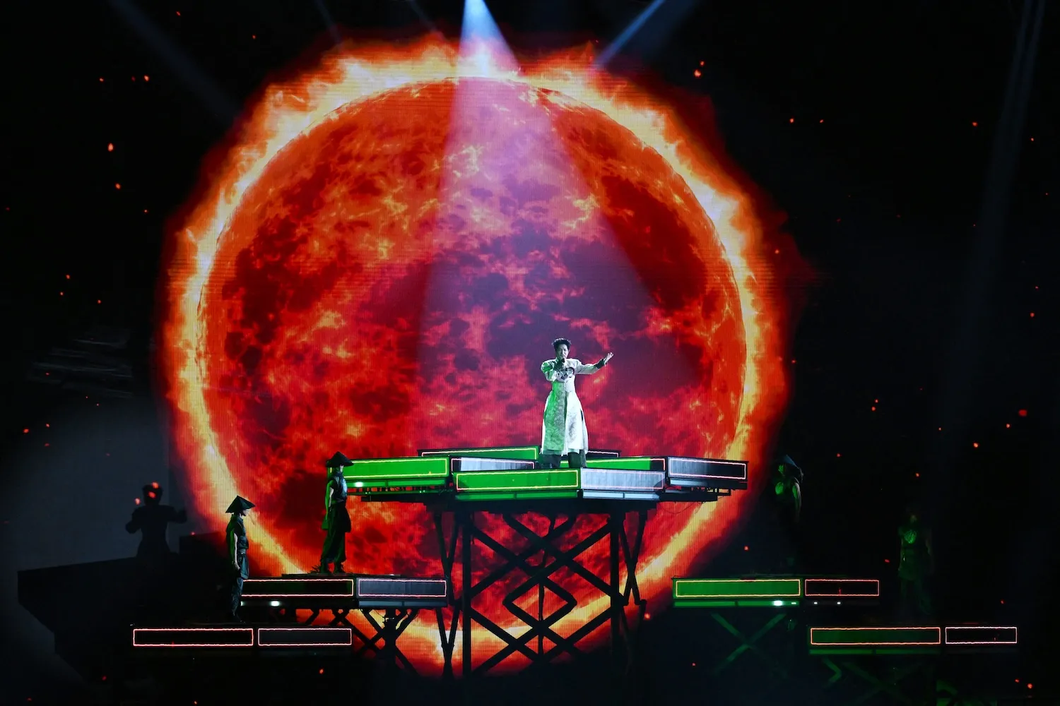 A singer stands on a green platform in front of a large picture of a burning planet.