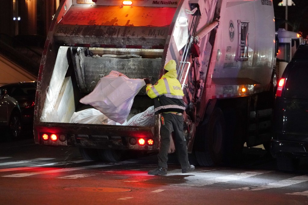 A DSNY Sanitation worker in a high-visibility jacket and hat loads garbage bags into the back of a garbage truck at night in New York City.
