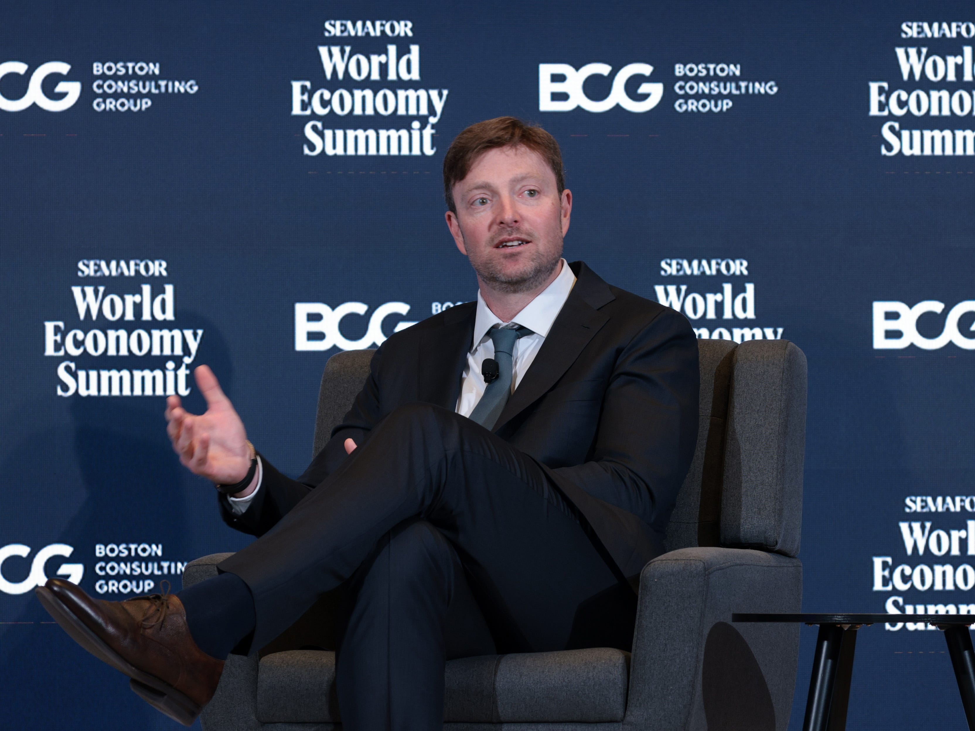 Alex Chriss wearing a suit with his legs crossed onstage.